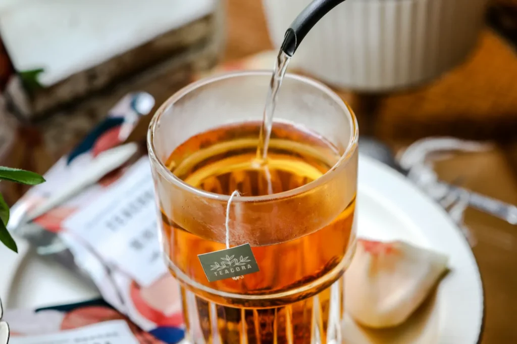 Close-up of a glass of amber tea being poured from a kettle, with a tea bag labeled "Teadora" inside. A delicate rose petal sits on the white saucer.