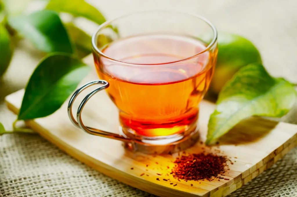 A clear glass cup of amber-colored tea on a wooden tray, surrounded by fresh green leaves and tea granules, creating a warm, natural ambiance.
