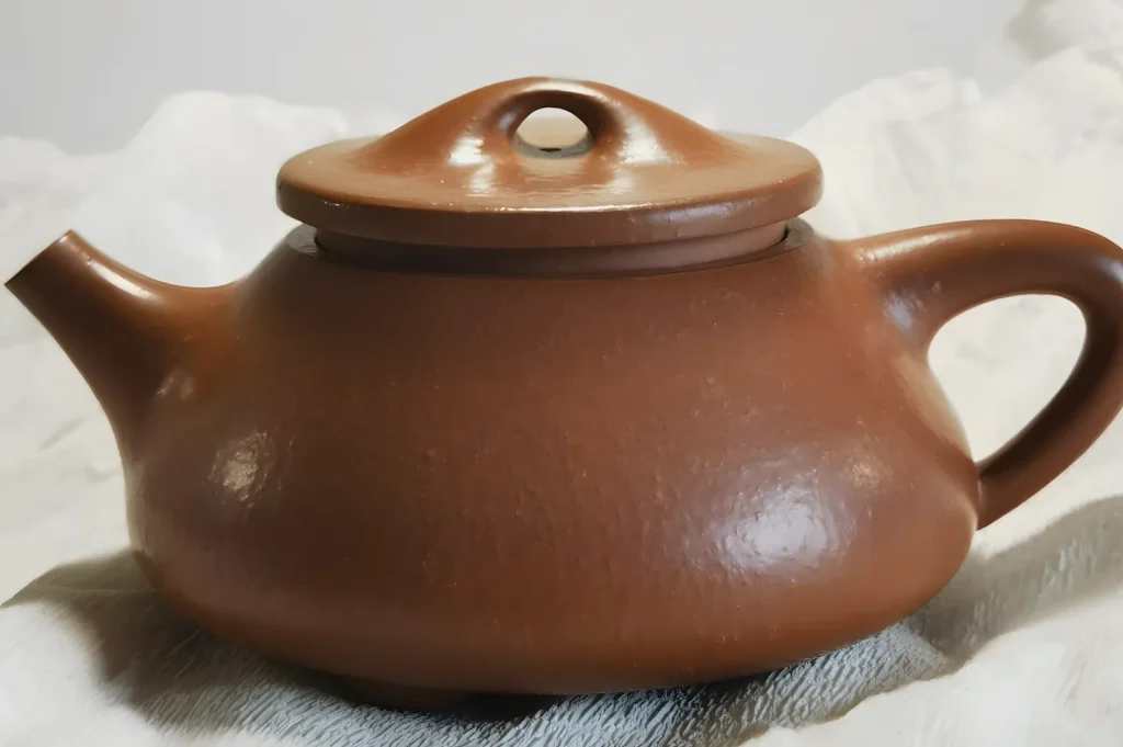 Brown ceramic teapot with a rounded body and lid, featuring a small spout and a curved handle. It rests on a soft, light-colored fabric.