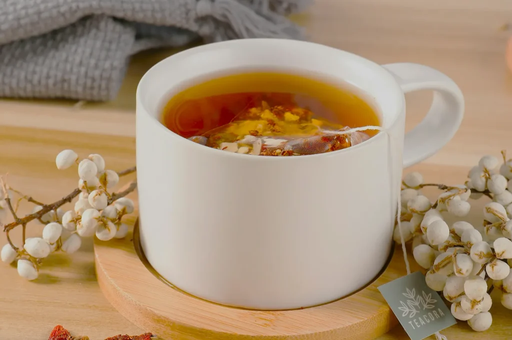 White mug of tea with a tea bag steeping inside, placed on a wooden coaster. Surrounding are white berries and a gray fabric, creating a cozy atmosphere.