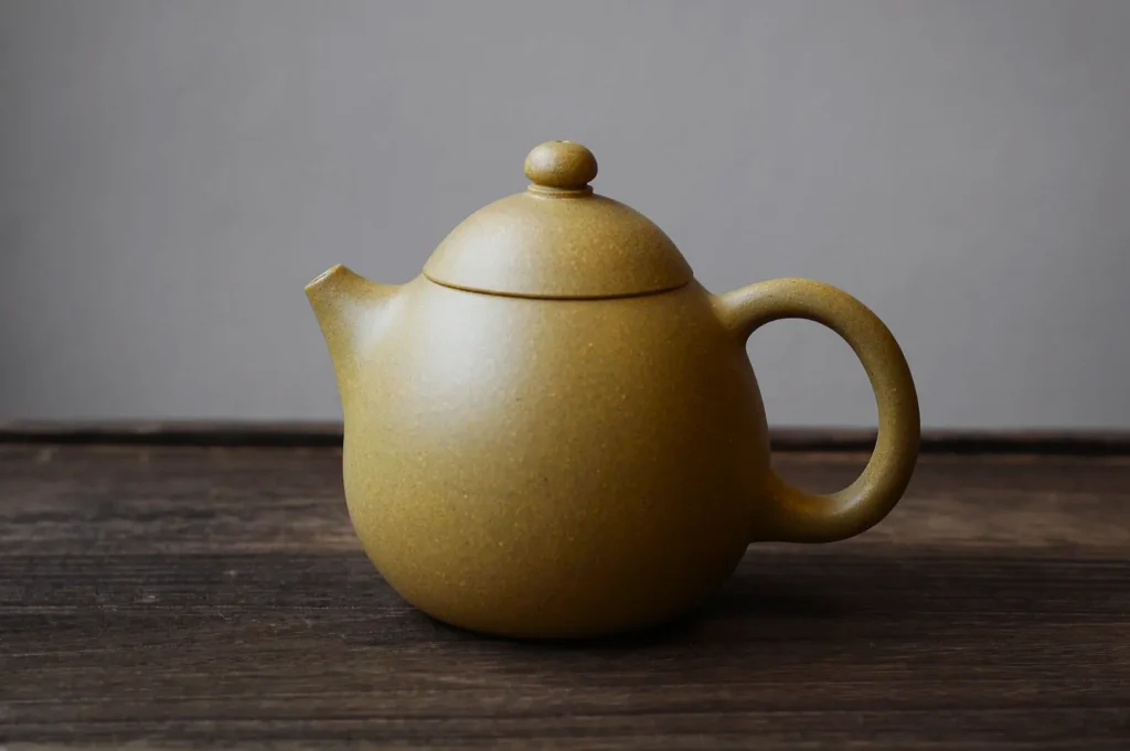 A simple, round, beige ceramic teapot with a curved handle and spout, sits on rustic wooden surface against a neutral gray background, conveying warmth.