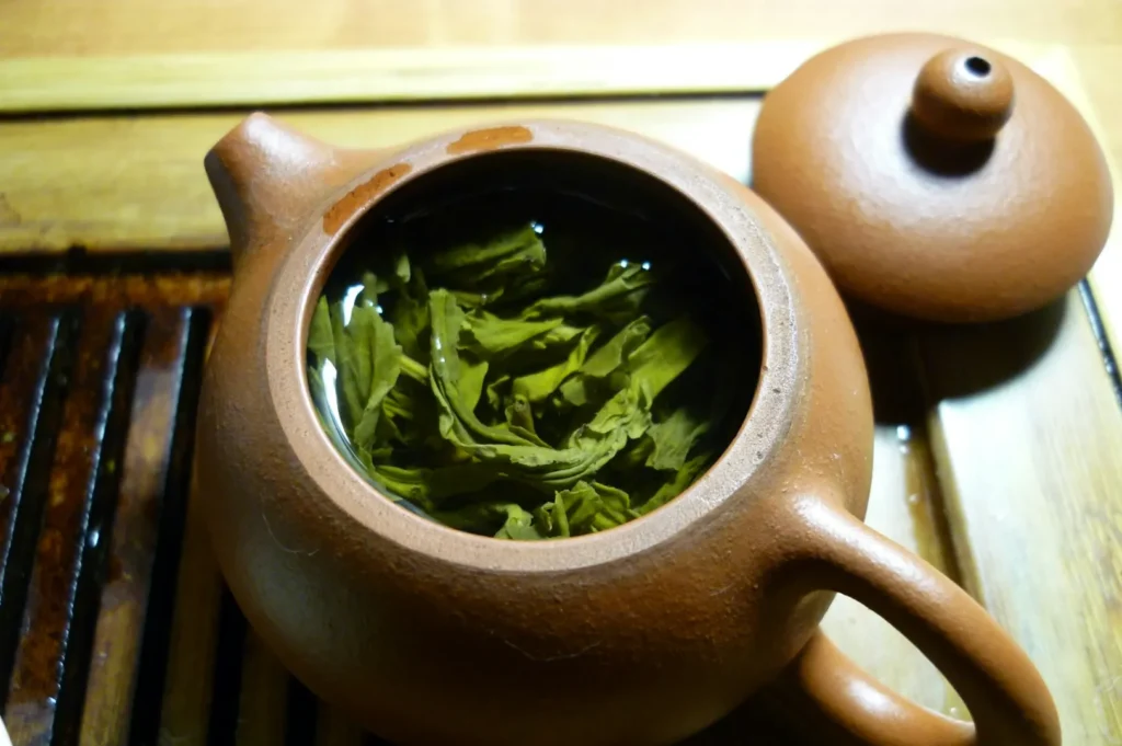 Close-up of a brown clay teapot with its lid off, revealing green tea leaves steeping in water. The warm wooden background adds a rustic feel.