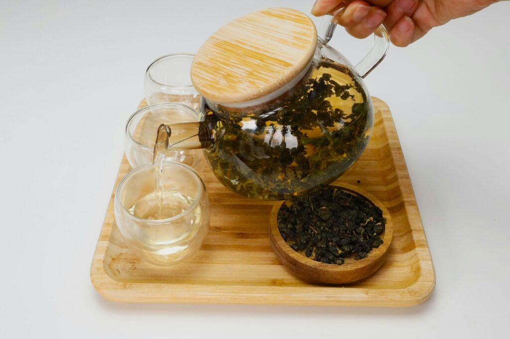 A hand pours tea from a glass teapot with a wooden lid into a clear glass cup, all placed on a bamboo tray with a small bowl of loose tea leaves.