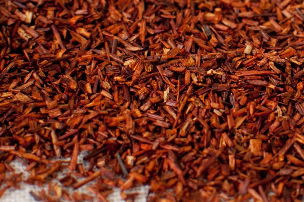 Close-up of loose rooibos tea leaves scattered on a white surface, displaying rich reddish-brown hues and a natural, rustic texture.