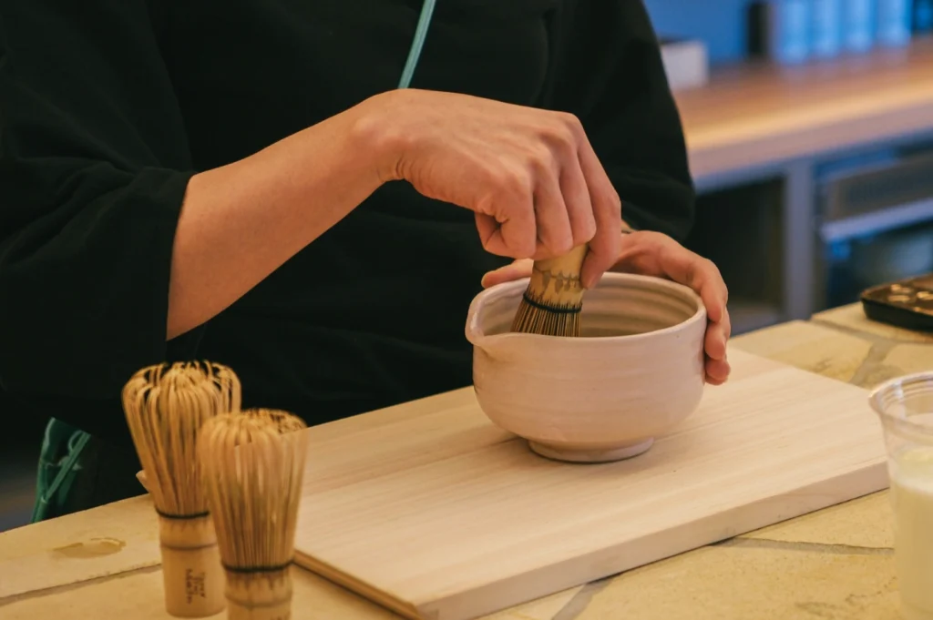 A person whisking matcha in a ceramic bowl with a bamboo whisk, over a wooden tray. The setting suggests a calm, focused tea preparation.