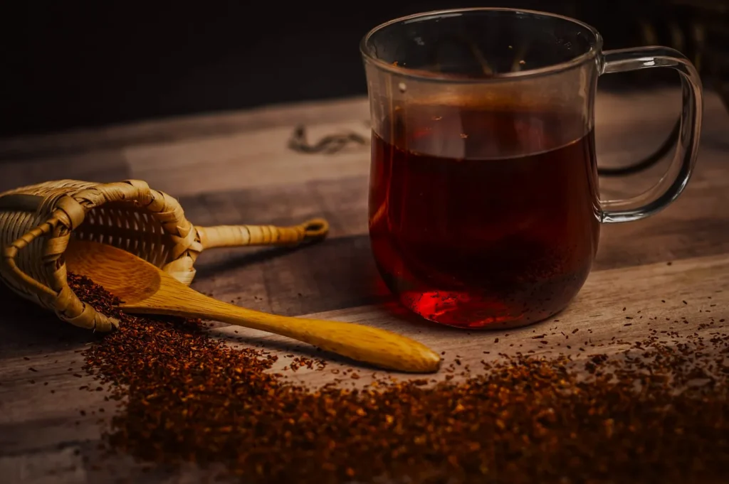 Glass mug of red rooibos tea on wooden table, beside a tipped basket, wooden spoon, and scattered loose tea, conveying warmth and rustic charm.