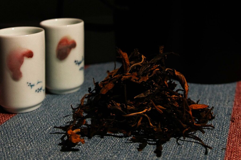 A rugged pile of dried tea leaves sits on a blue woven mat beside two white porcelain cups featuring subtle pink motifs, against a dark background at Tea Room by Ki-setsu.