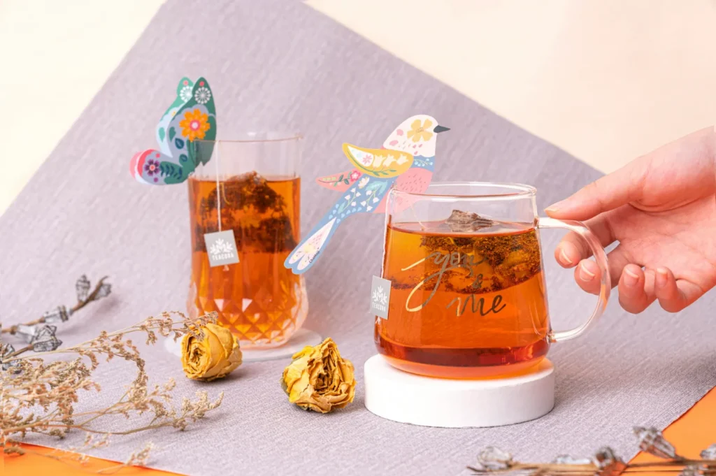 Two glasses of amber tea with whimsical bird-shaped tags sit on coasters. A hand holds one mug, surrounded by dried flowers on a gray cloth.