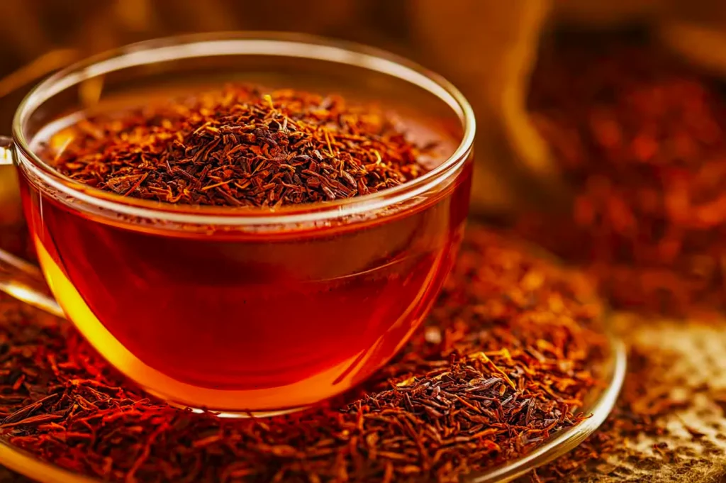 A clear glass teacup filled with rich amber tea is topped with a mound of dry rooibos leaves. More leaves are scattered on a rustic wooden surface.