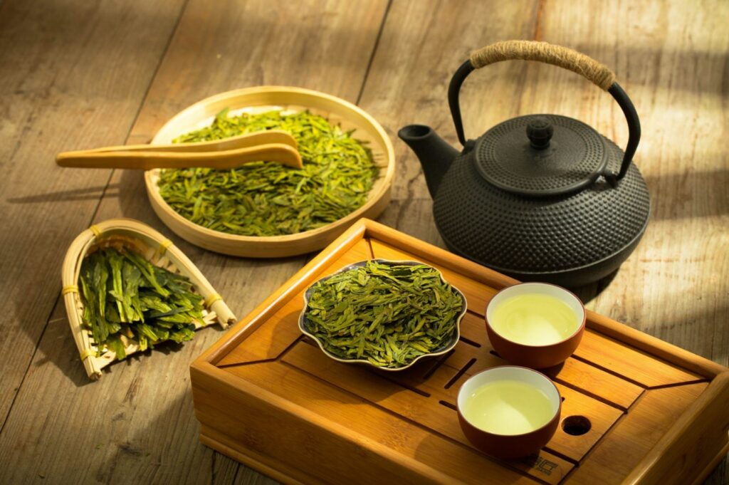 Close view of green tea leaves spread in a shallow wooden tray and a wooden scoop resting across them, while a dark cast-iron kettle sits in the background.