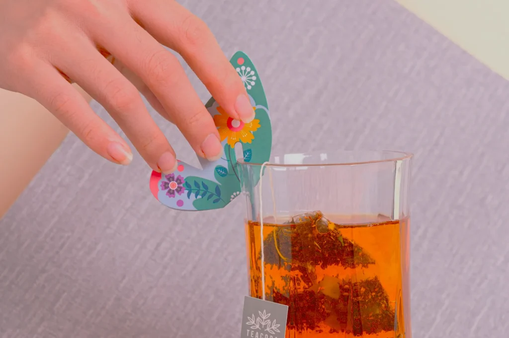 A hand holds a colorful, floral-patterned butterfly decoration above a glass of amber tea with a tea bag inside, conveying a serene, cozy vibe.