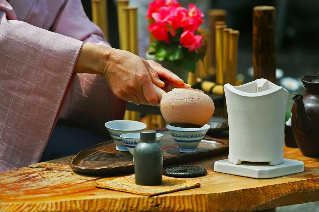 A hand gracefully handles a traditional tea ladle over porcelain cups on a wooden table. Pink flowers and bamboo in the background add elegance.