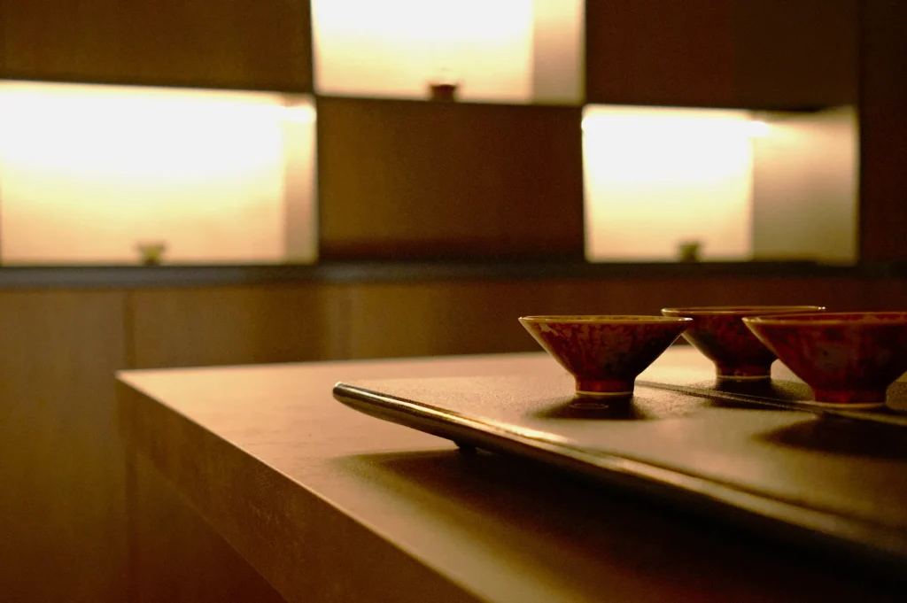 A serene, minimalist setting with dim lighting. Four small ceramic bowls sit on a tray in the foreground, while softly lit shelves create a warm, calm ambiance.