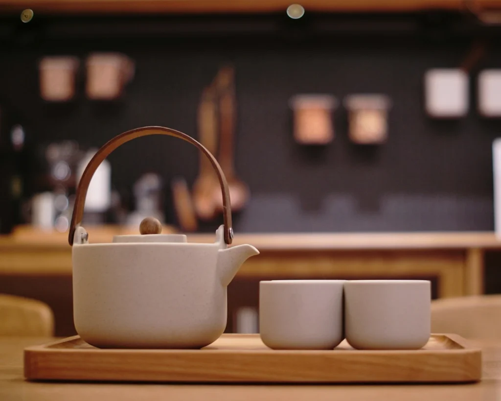Ceramic teapot with a wooden handle and two cups on a wooden tray, set against a softly blurred kitchen backdrop creating a cozy, inviting atmosphere.
