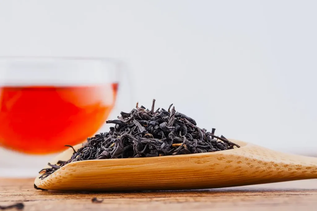 Loose black tea leaves on a wooden spoon, with a glass cup of brewed tea in the background. The scene conveys warmth and relaxation.