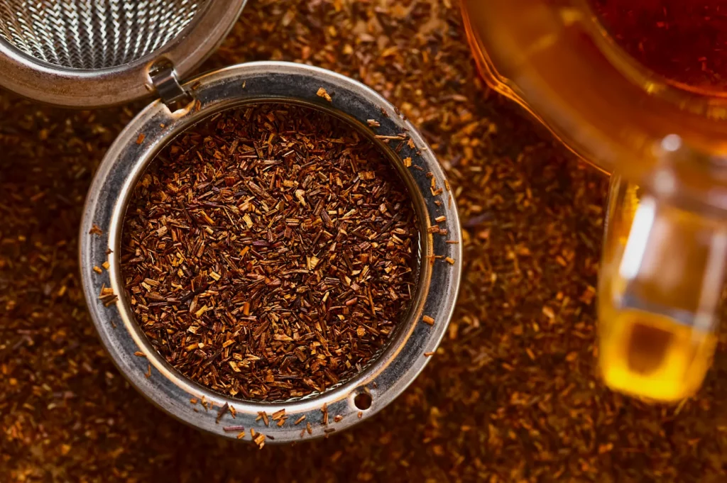A close-up of an open tea infuser filled with brown rooibos tea leaves on a matching background. A glass teapot with brewed tea is visible on the right.