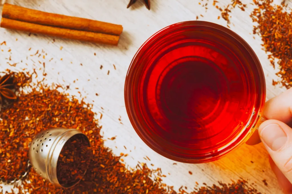 A hand holds a warm glass of red rooibos tea on a light wooden table. Loose tea leaves, a tea infuser, and cinnamon sticks are scattered around, creating a cozy atmosphere.