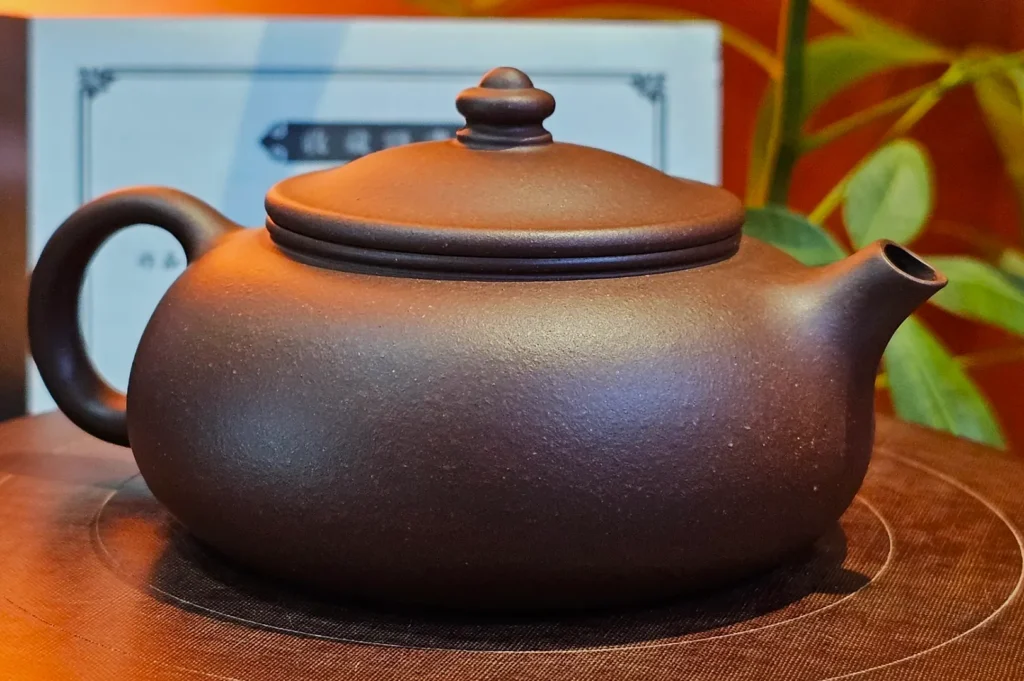 A round, brown clay teapot with a short spout and curved handle is displayed on a mat. In the background, there are blurred plants and a framed sign.