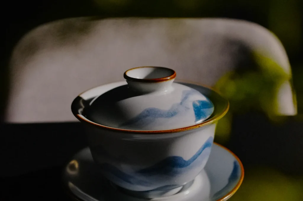 A ceramic teacup with a lid and saucer, featuring a soft blue wave design, sits in gentle light on a dark surface, creating a serene, elegant ambiance.