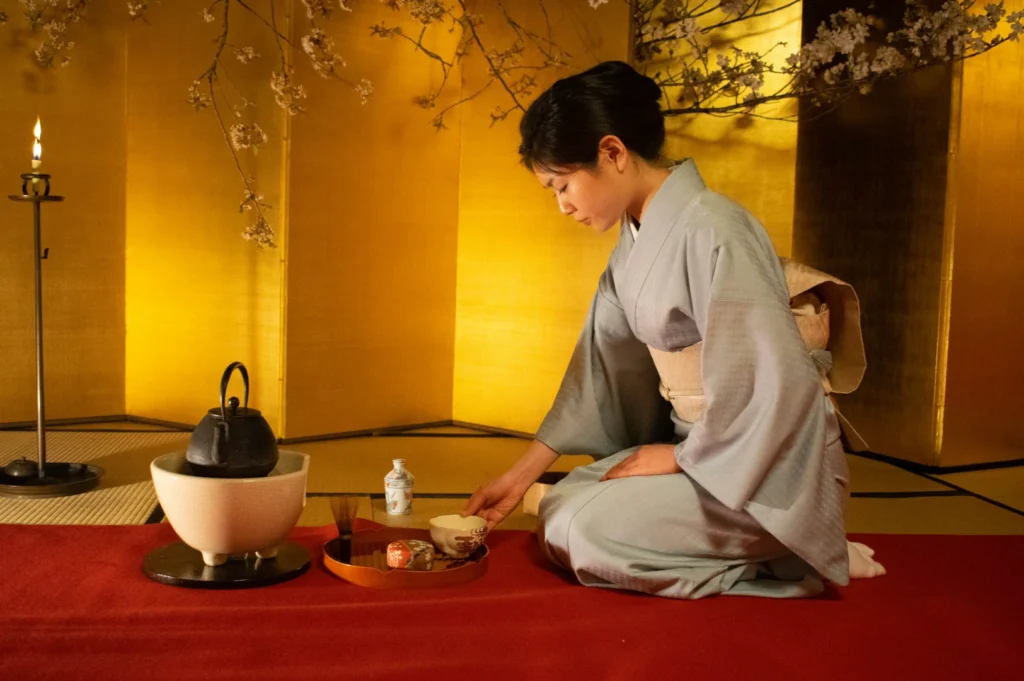 A woman in a kimono performs a traditional Japanese tea ceremony. She kneels on a red mat, surrounded by a serene, gold-lit room with cherry blossoms.