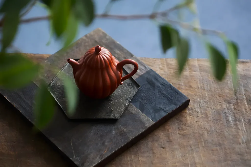 A small, ribbed terracotta teapot sits on layered slate tiles on a wooden surface. Green leaves blur in the foreground, adding a serene touch.