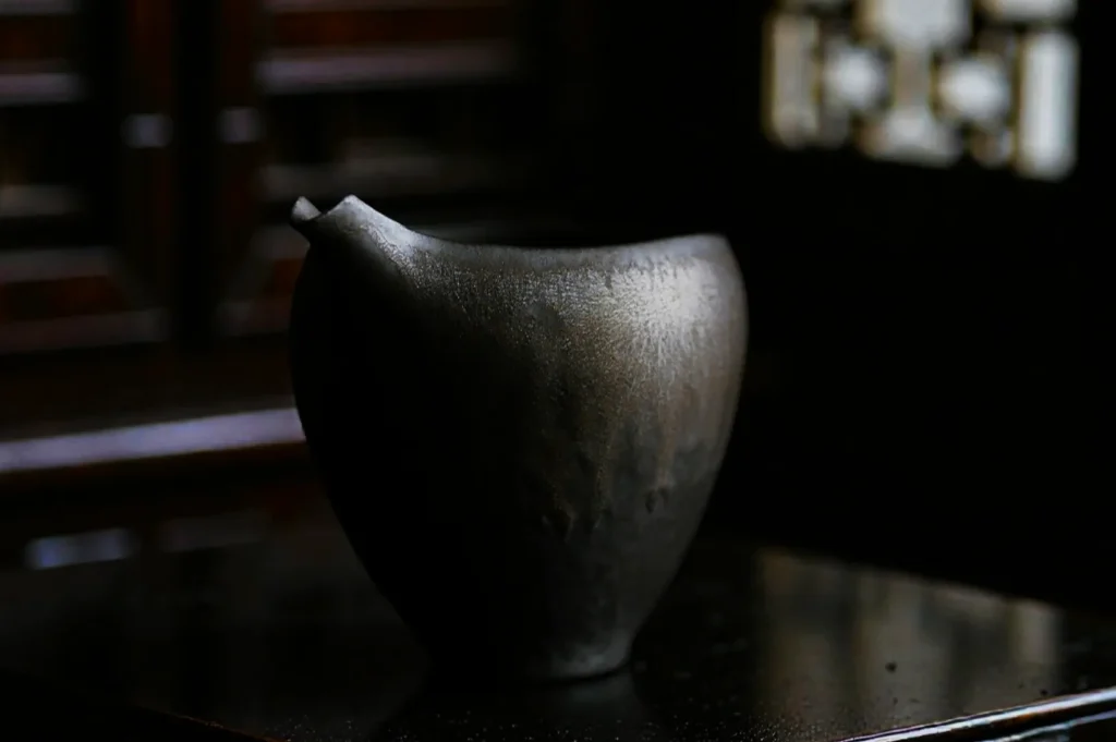 A dark ceramic pitcher with a small spout sits on a reflective surface in a dimly lit room, creating a moody and contemplative atmosphere.