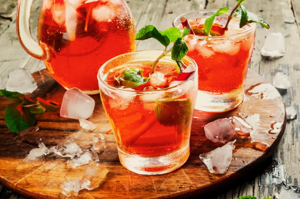 Close-up of two glasses and a jug of iced red tea garnished with fresh mint on a wooden board, surrounded by ice cubes, creating a refreshing and inviting scene.