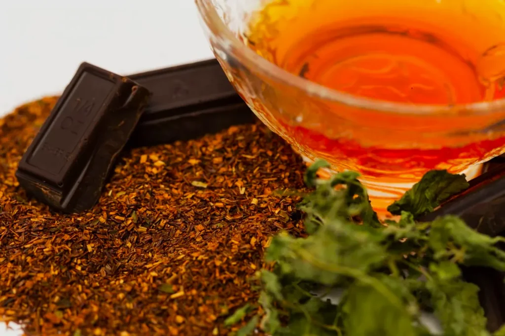 A glass teacup with amber-colored tea sits next to dark chocolate pieces, loose rooibos tea leaves, and fresh mint, creating a warm and cozy atmosphere.