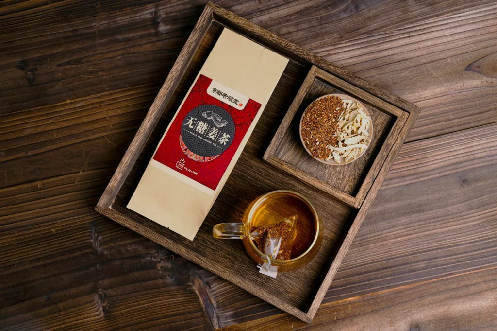 A wooden tray holds a tea package, a glass cup of brewed tea with a tea bag, and a small bowl of loose leaf tea. The setting is warm and inviting.