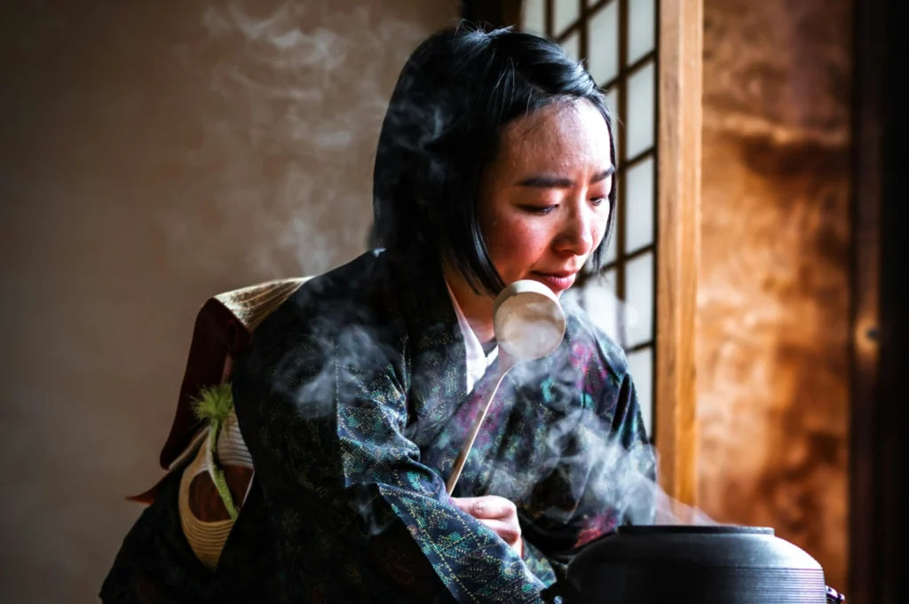 A person in traditional attire performs a tea ceremony, gracefully pouring hot water from a ladle, with steam rising in a serene, softly lit room.