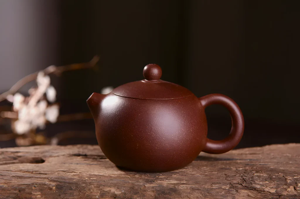 A smooth, round brown teapot with a short spout and curved handle sits on a rustic wooden surface. Blurred white flowers are in the background. The scene feels serene and timeless.