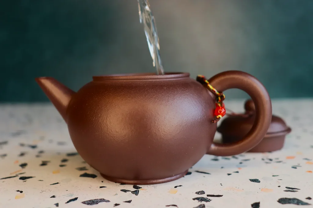 A brown teapot with a curved handle and small charm is being filled with water. Its lid is off and rests on a speckled countertop. The scene is calm.