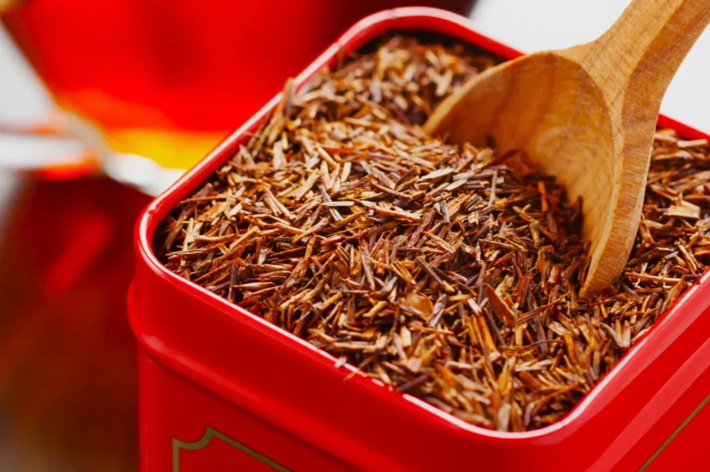 Close-up of a red square tin filled with loose rooibos tea leaves, with a wooden spoon on one side. A warm, cozy atmosphere is conveyed.