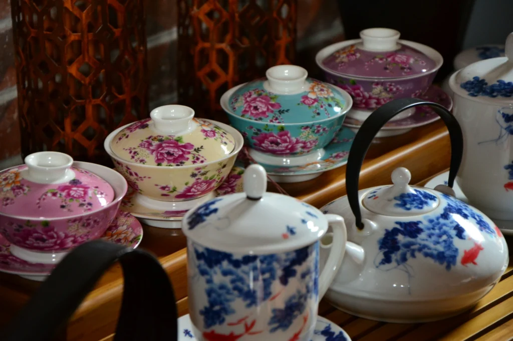 Assorted teacups with floral designs sit on a wooden tray. A blue and white teapot with a black handle is in the foreground, conveying a cozy, elegant atmosphere.