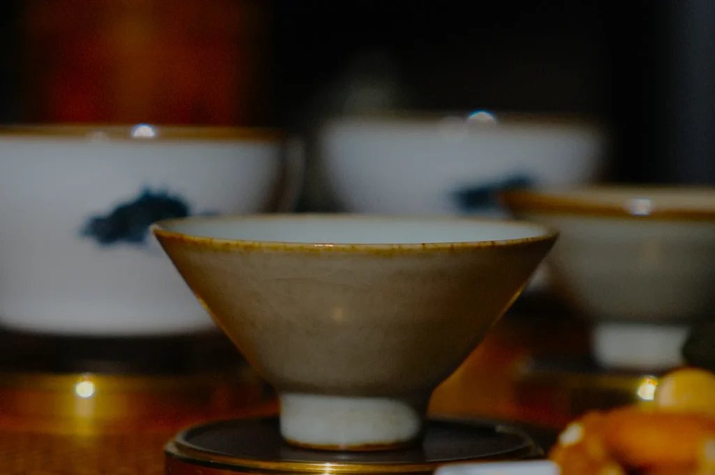 Close-up of a rustic ceramic bowl on a wooden table, surrounded by blurred white cups with blue patterns, creating a warm and cozy ambiance.