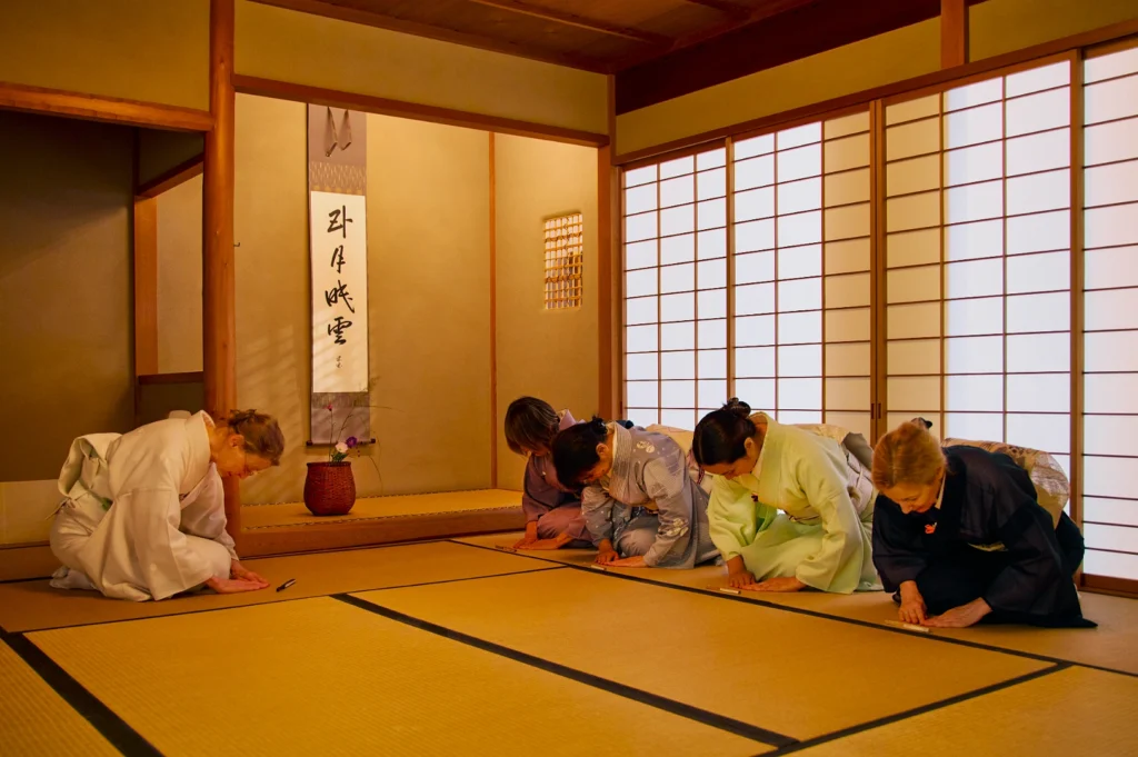 Five individuals in traditional Japanese attire perform a formal bow in a serene tea room, featuring tatami mats, shoji screens, and a calligraphy scroll.