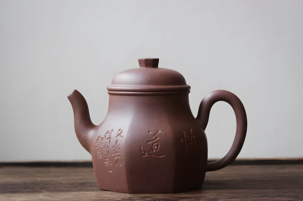 Brown clay teapot with intricate Chinese characters, short spout, and curved handle on wooden surface. Minimalist, serene, and traditional design.