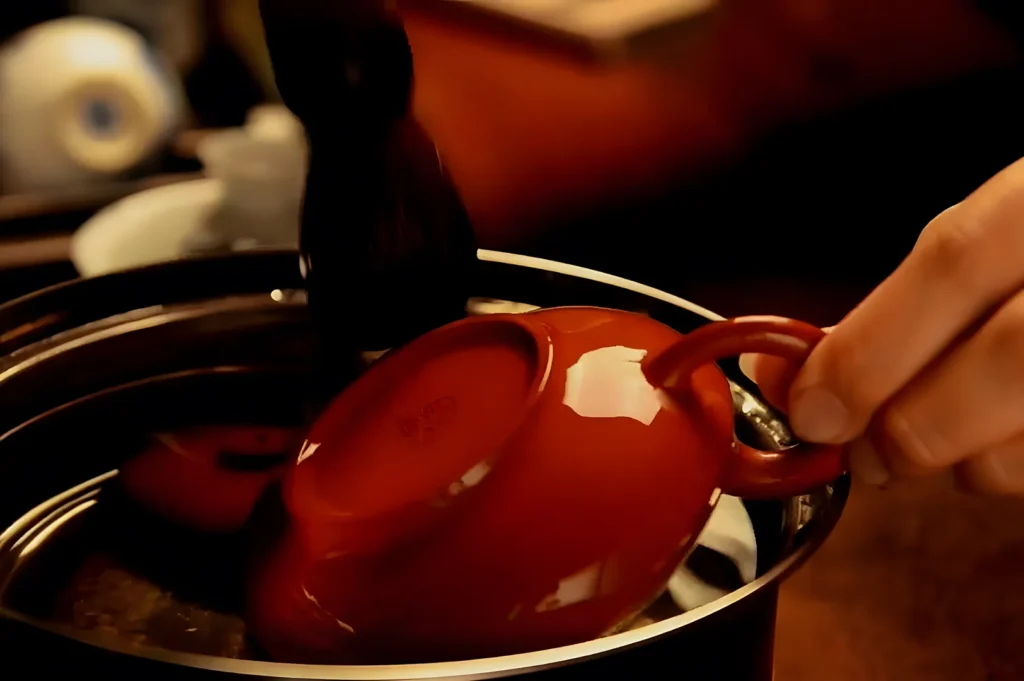 A hand gently holds a red ceramic cup over a pot, engaging in a tea ceremony. The warm lighting conveys a sense of calm and tradition.