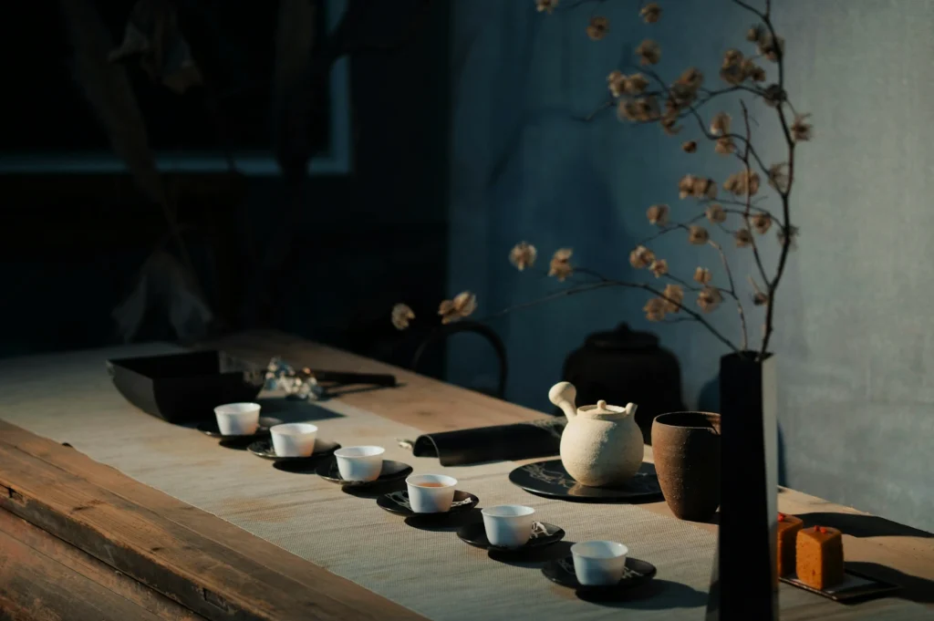 Minimalist tea setting on a wooden table with a white teapot, six small cups on black saucers, and a tall vase holding delicate branches, creating a serene atmosphere.