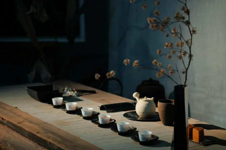 Minimalist tea setting on a wooden table with a white teapot, six small cups on black saucers, and a tall vase holding delicate branches, creating a serene atmosphere.