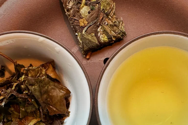 Two ceramic bowls on a brown tray. One holds brewed tea leaves, the other has yellow tea liquid. A compressed tea block is above the bowls.