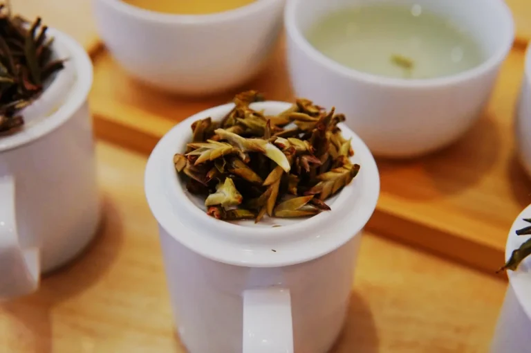 White teacup with steeped tea leaves on lid, surrounded by two bowls of light-colored tea on a wooden tray, conveying a calm, aromatic setting