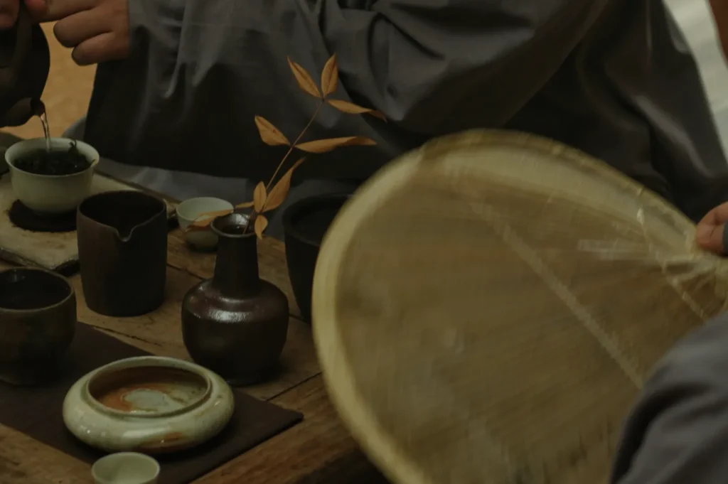 Hands prepare tea in a serene setting, surrounded by rustic pottery, cups, and a woven fan. A branch with brown leaves adds an autumnal touch.