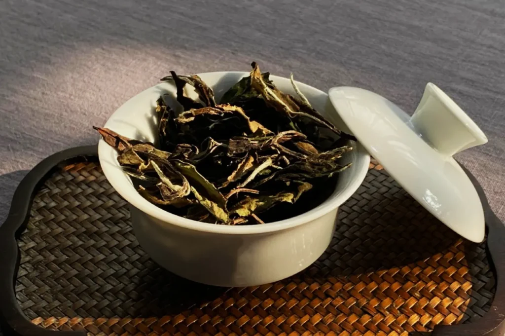 A white ceramic bowl filled with dried tea leaves sits on a woven mat. The lid is slightly open, and soft sunlight casts a gentle shadow.