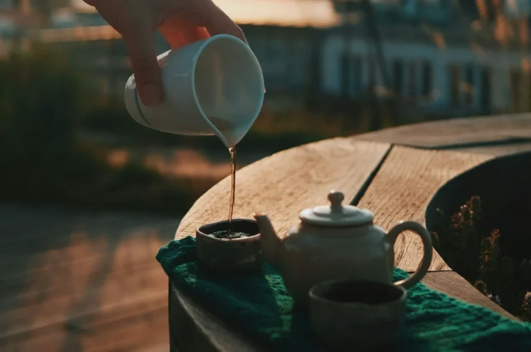 A hand pours tea from a white cup into a small ceramic teapot on a wooden table, with a green cloth under the teapot. The scene is warmly lit by the sunset.