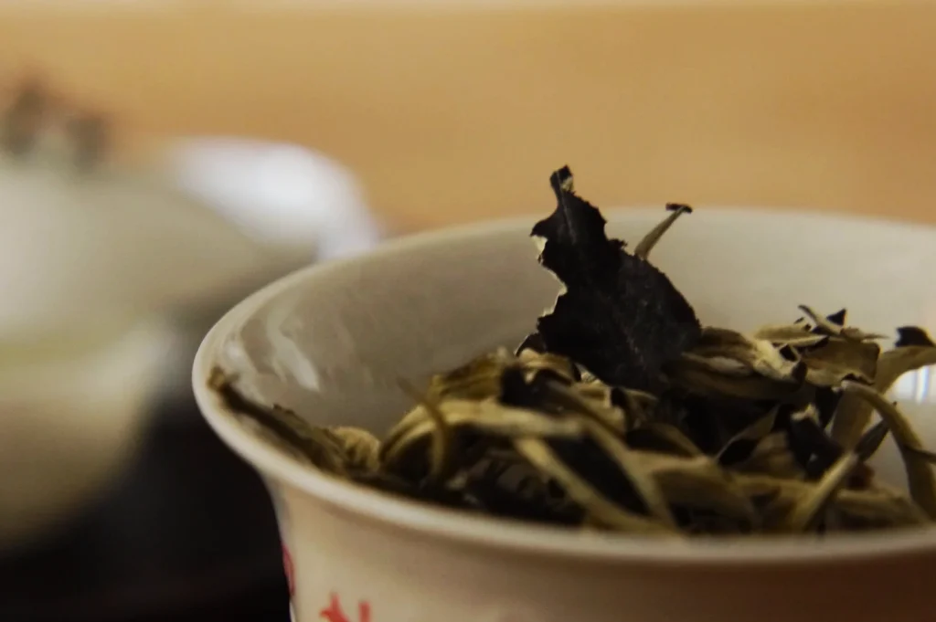 A close-up of a white bowl containing dried tea leaves. The focus highlights the texture and dark color of the leaves against a soft, blurred background.