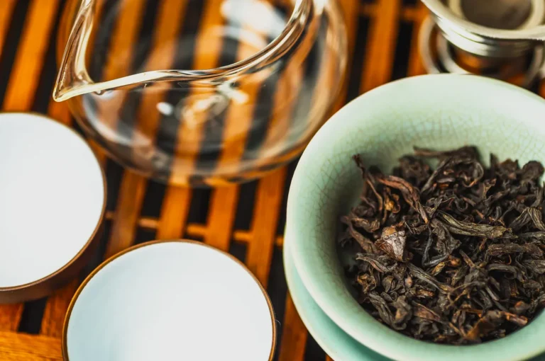 A close-up of a wooden tray with a glass teapot, two white teacups, and a green bowl filled with loose dark tea leaves, conveying a calm tea-making setup.