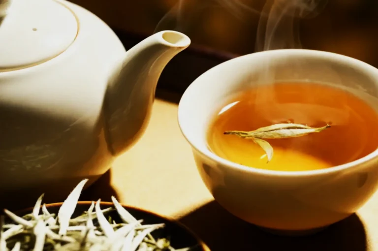 A steaming cup of amber tea with a floating leaf sits beside a white teapot. Green tea leaves are visible in the foreground, evoking a calming ambiance.