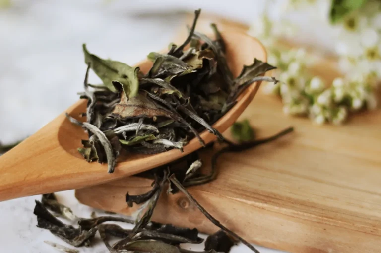 Dried tea leaves on a wooden spoon rest on a wooden board. Soft focus on small white flowers in the background, creating a calm, natural feel.