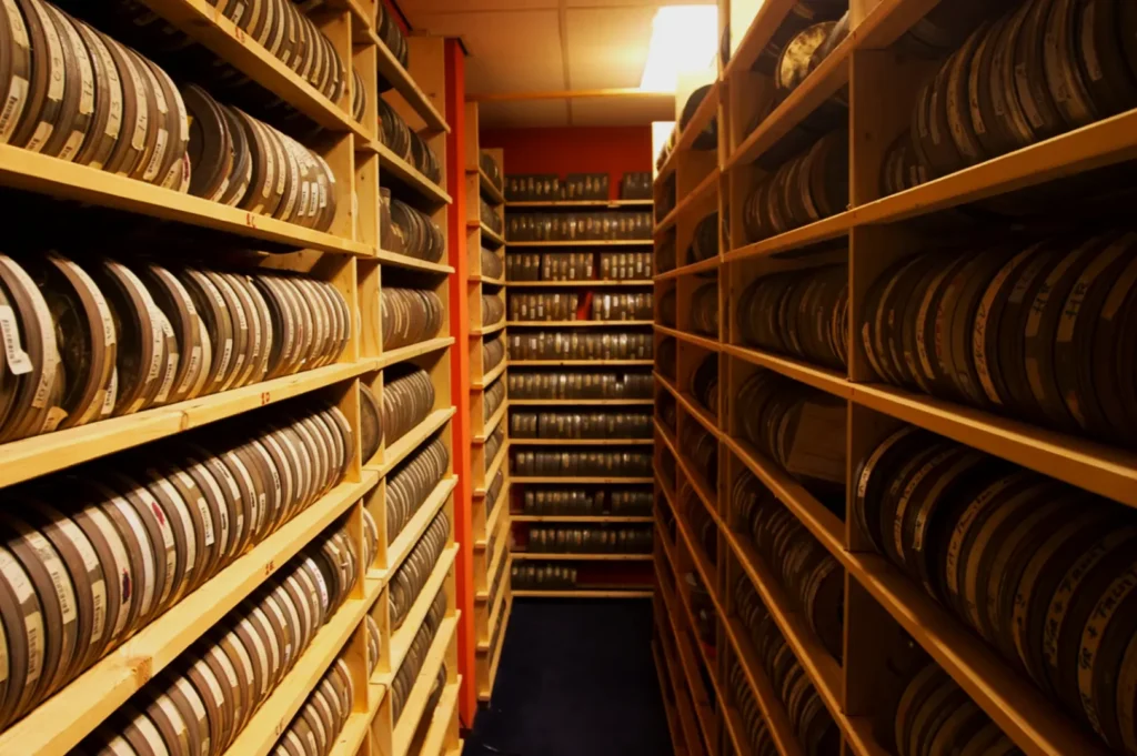Dimly lit archive room filled with shelves lined with film reels. The narrow aisle emphasizes a sense of nostalgia and preservation.